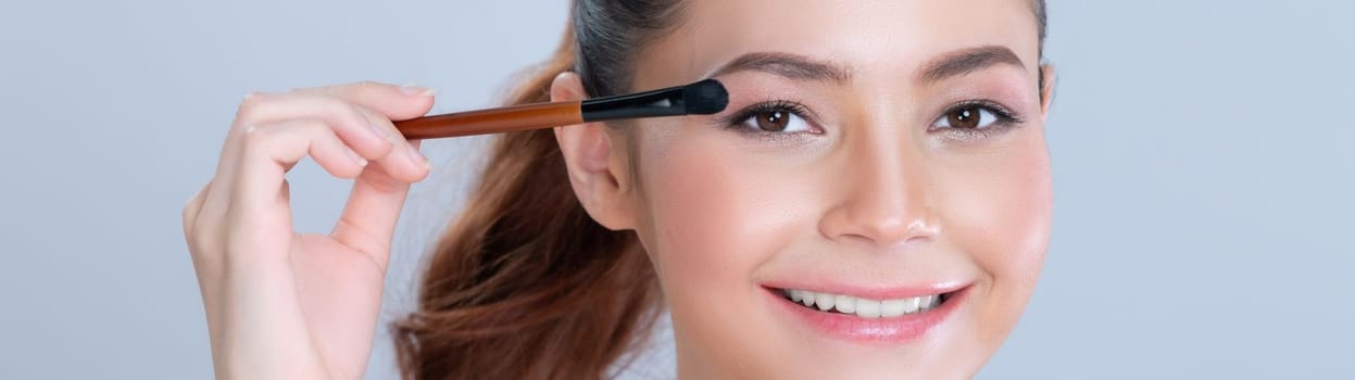 Closeup beautiful girl with flawless applying glamorous eye shadow makeup with eyeliner brush. Cosmetic facial painting process on lovely young woman with perfect clean skin in isolated background.