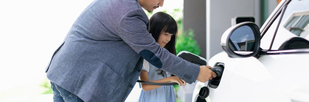 Progressive father and daughter plugs EV charger from home charging station to electric vehicle. Future eco-friendly car with EV cars powered by renewable source of clean energy.