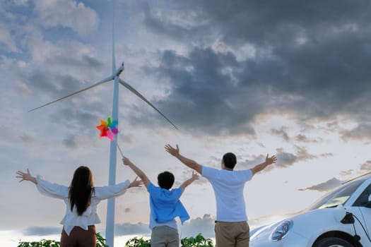 Concept of progressive happy family enjoying their time at wind farm with electric vehicle. Electric vehicle driven by clean renewable energy from wind turbine generator for charging station.