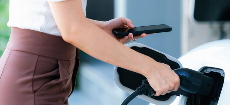 Closeup progressive woman install cable plug to her electric car with home charging station. Concept of the use of electric vehicles in a progressive lifestyle contributes to clean environment.