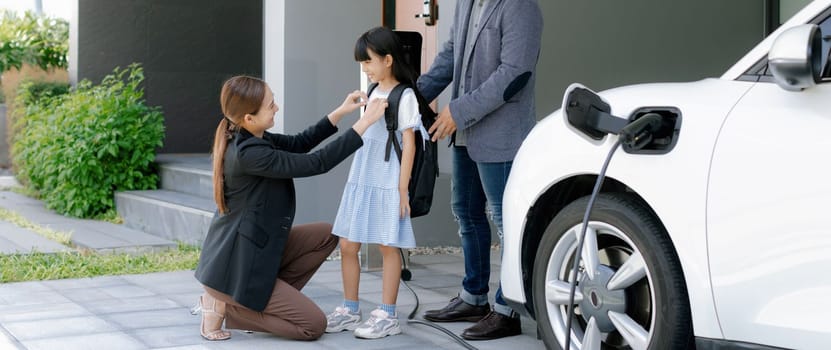 Progressive young parents and daughter with electric vehicle and home charging station. Green and clean energy from electric vehicles for healthy environment. Eco power from renewable source at home.