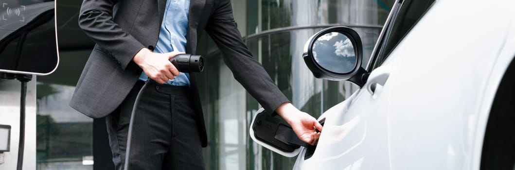 Closeup progressive black suit businessman insert EV charger plug from public charging station to his electric car. Eco friendly electric rechargeable car with zero CO2 emission.