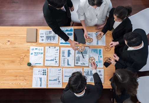Top view hand holding gear by businesspeople wearing suit for harmony synergy in office workplace concept. Group of people hand making chain of gears into collective form with dashboard report papers.