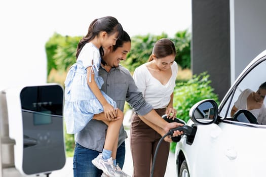 Progressive young parent teach daughter how to recharge or refuel EV car at home charging station. Green and clean energy from electric vehicle for healthy environment. Eco power from renewable source