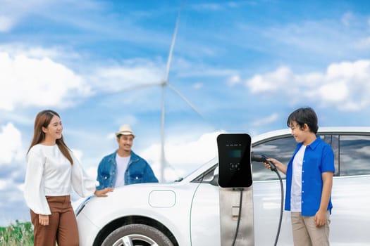 Concept of progressive happy family enjoying their time at wind farm with electric vehicle. Electric vehicle driven by clean renewable energy from wind turbine generator for charging station.