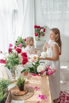 A little blonde girl with her mom on a kitchen countertop decorated with peonies. The concept of the relationship between mother and daughter. Spring atmosphere.