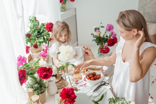 A little blonde girl with her mom on a kitchen countertop decorated with peonies. The concept of the relationship between mother and daughter. Spring atmosphere.