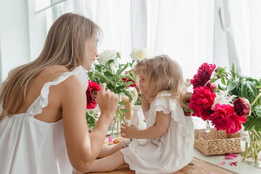 A little blonde girl with her mom on a kitchen countertop decorated with peonies. The concept of the relationship between mother and daughter. Spring atmosphere.