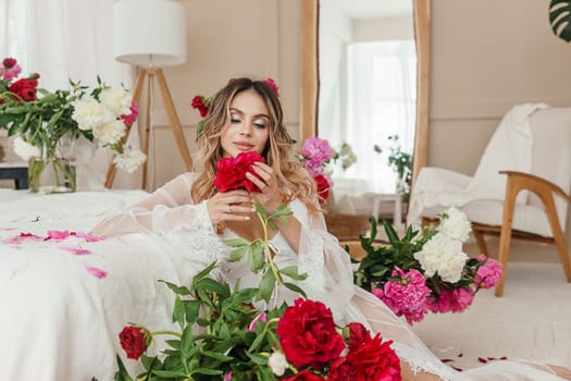 A beautiful girl in a white nightgown at home, surrounded by spring flowers on a large bed. A room decorated with bouquets of peonies.
