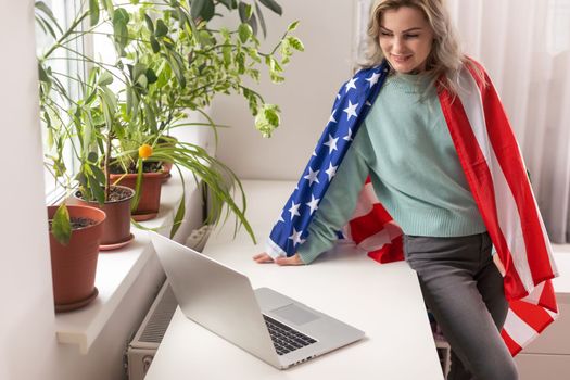 Happy woman employee sitting wrapped in USA flag, shouting for joy in office workplace, celebrating labor day or US Independence day