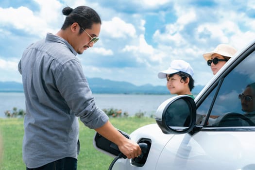 Concept of progressive happy family with electric vehicle enjoy their time at natural outdoor scenic with lake, greenfield and cloudscape background. Electric vehicle driven by green renewable energy.