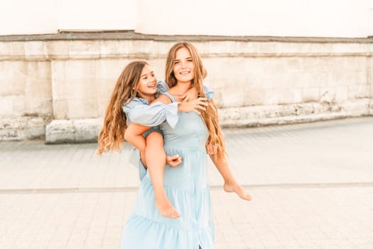 Mother of the daughter walks playing. Mother holds the girl on her back, holding her legs, and her daughter hugs her by the shoulders. Dressed in blue dresses