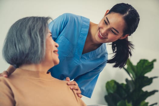 A caregiver rest her hands on the shoulders of a contented senior patient while she sitting on the sofa at home.