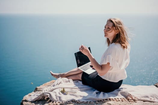 Successful business woman in yellow hat working on laptop by the sea. Pretty lady typing on computer at summer day outdoors. Freelance, travel and holidays concept.