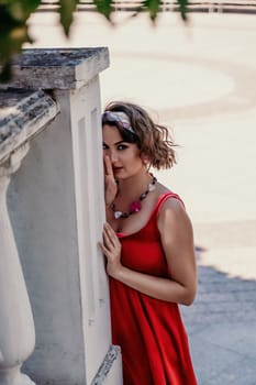 woman in a red silk dress and a bandage on her head smiles against the background of the leaves of a tree. She is leaning on the coop and looking into the camera. Vertical photo