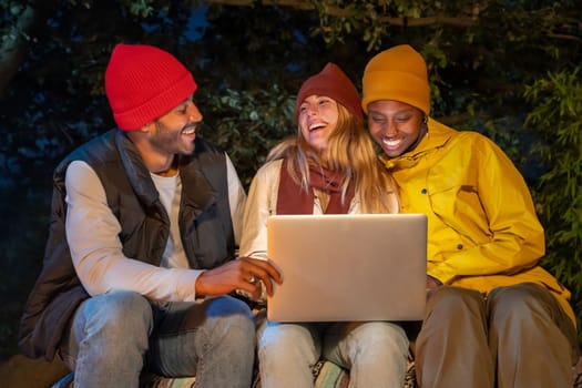 Group of interracial young people using laptop while camping in countryside at night, video chatting online, having fun, laughing.
