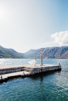 Bride in a flowing dress stands on a pier near a column with a cross. High quality photo