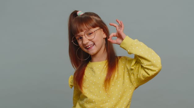 Ok. Young toddler school girl in glasses looking approvingly at camera showing okey gesture, like sign positive, approve something good. Teenager female child kid isolated on studio gray background