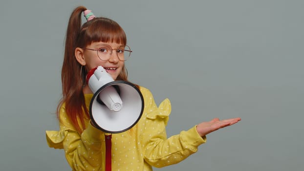 Young toddler girl in glasses scream shout in megaphone loudspeaker announces discounts sale, announcing advertisement, pointing empty place area. Preteen child kid isolated on studio gray background