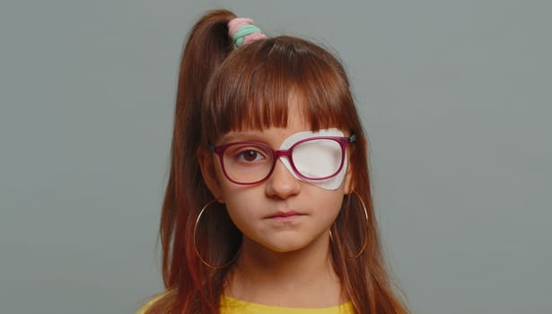 Sick ill preteen girl looking at camera having protective bandage plaster patch scratch on one eye. Injury, trauma, blind. Child kid wearing glasses at gray background. Medical eyesight vision therapy