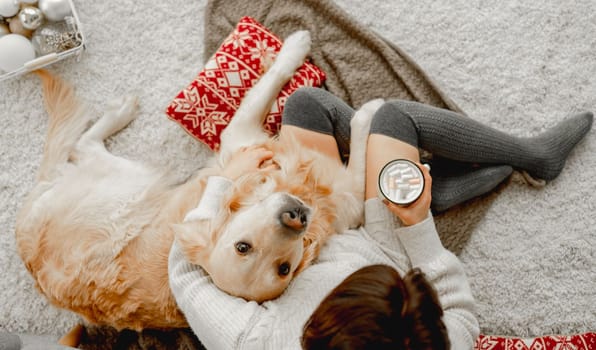 Girl holding mug with cocoa and marshmallow and golden retriever dog looking at camera in festive holidays time. Young woman with hot beverage and pet doggy at home together