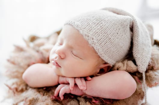 Newborn baby child wearing hat sleeping and holding hands under cheeks. Sweet infant kid napping closeup portrait