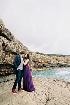Woman put her hand on the chest of man hugging her waist on the rocks by the sea. High quality photo