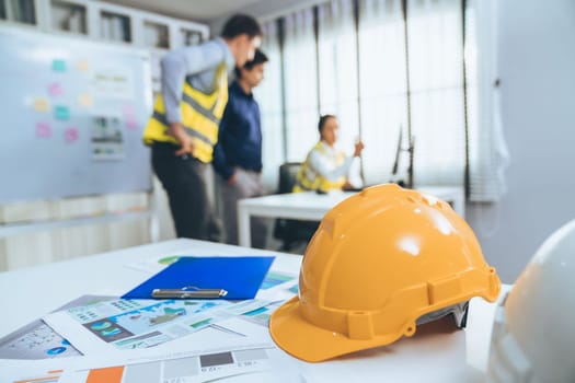 Concept of engineering equipments of protective helmet for competent engineer on piles of paper works. Blurred image of engineers and employer working together at the desk.