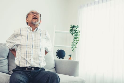 An agonizing senior man in need of assistance while sitting on his sofa at home, suffering from back pain. Senior care, nursing home for pensioners, deteriorating health of old age.