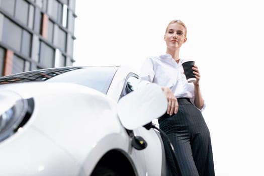 Businesswoman drinking coffee, leaning on electric vehicle recharging at public charging station with residential apartment condos building in background as progressive lifestyle by eco-friendly car.