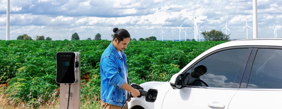 Progressive man with his electric car, EV car recharging energy from charging station on green field with wind turbine as concept of future sustainable energy. Electric vehicle with energy generator.