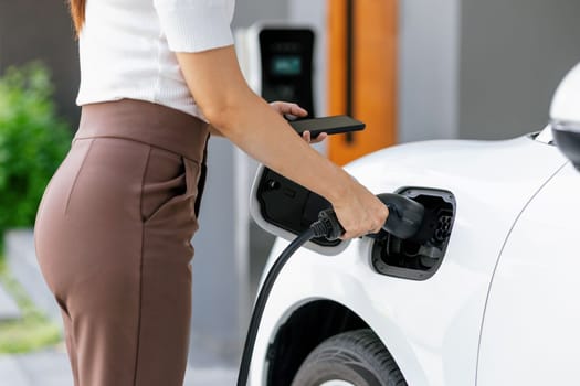 Closeup progressive woman install cable plug to her electric car with home charging station. Concept of the use of electric vehicles in a progressive lifestyle contributes to clean environment.