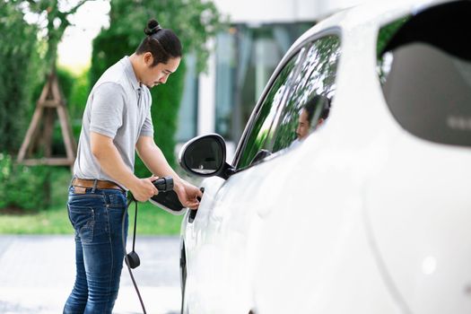 Progressive asian man install cable plug to his electric car with home charging station in the backyard. Concept use of electric vehicles in a progressive lifestyle contributes to clean environment.