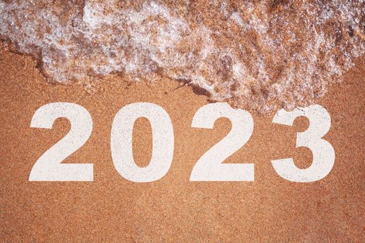 The concept of plans for 2023. A wave of water washes away the figure 2023 on the sand on the seashore or ocean. Year 2023 handwriting on sand beach surface.