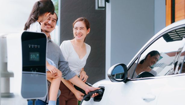 Progressive young parents and daughter with electric vehicle and home charging station. Green and clean energy from electric vehicles for healthy environment. Eco power from renewable source at home.