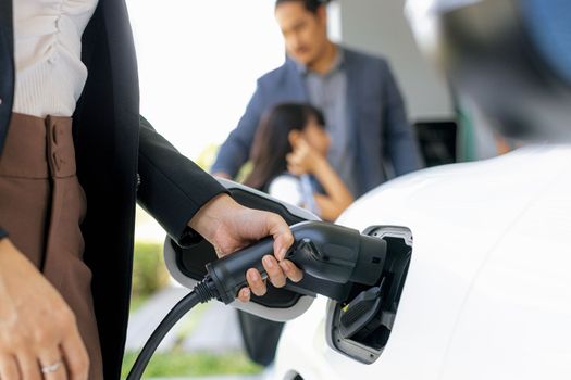 Focus closeup hand charging electric car, insert charger device into electric vehicle as progressive lifestyle concept of alternative green energy technology with blurred family in the background.