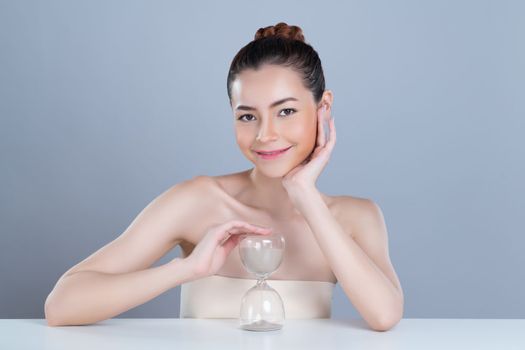 Glamorous model holding hourglass in beauty concept of anti-aging skincare treatment for woman. Young asian girl portrait with perfect smooth clean skin and flawless soft makeup in isolated background