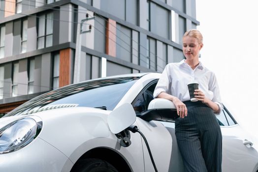 Businesswoman drinking coffee, leaning on electric vehicle recharging at public charging station with residential apartment condos building in background as progressive lifestyle by eco-friendly car.