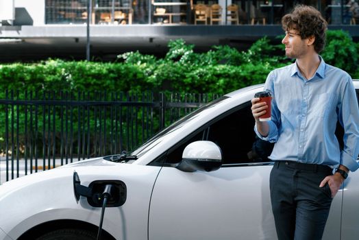 Progressive eco-friendly concept of parking EV car at public electric-powered charging station in city with blur background of businessman leaning on recharging-electric vehicle with coffee.