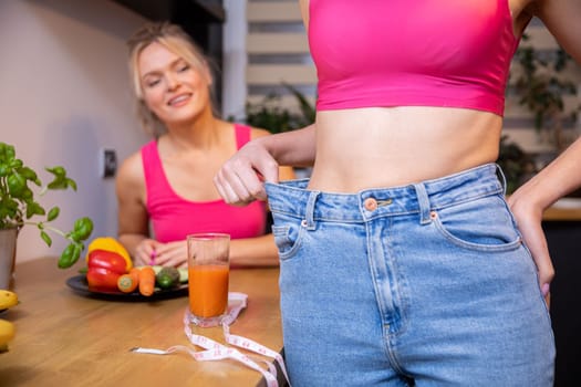 Friends at a kitchen meeting look at the flat belly after weight loss of one of them. On the table are plates of fruits and vegetables and a glass of juice wrapped in measure tape.