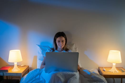 Beautiful young girl sitting on bed at night working with her laptop in the middle of the night. High quality photo