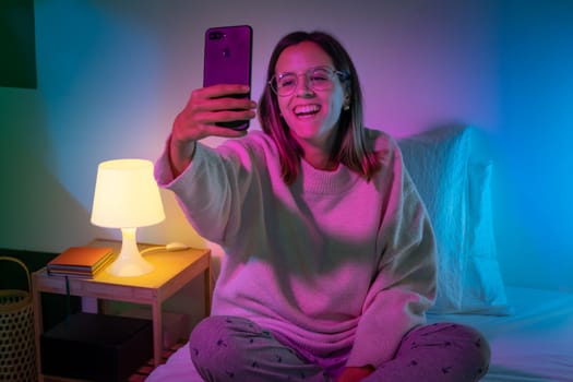 Beautiful young girl taking selfie with her phone on bed with neon colors room. Technology at bed concept. High quality photo