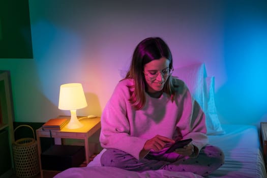 Beautiful young girl looking her phone on bed with neon colors room. Technology at bed concept. High quality photo