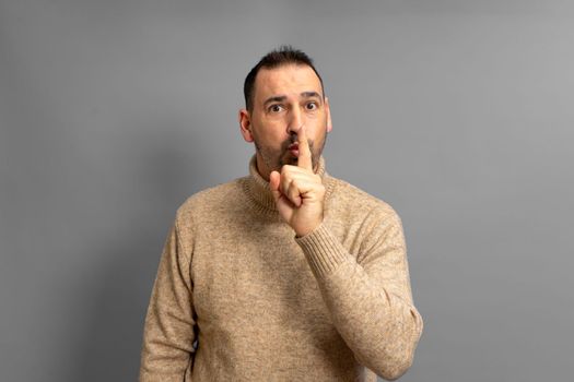 Hispanic handsome man wearing beige turtleneck over isolated gray background asking for silence with finger on lips. Silence and secret concept.