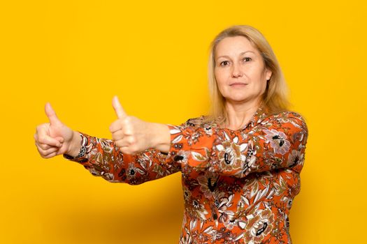 Caucasian woman in her 40s wearing a patterned dress showing thumbs up in agreement, isolated over yellow background