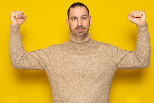 Bearded hispanic man wearing a beige turtleneck showing his biceps isolated over yellow background, he is in lousy shape due to sloppiness and sedentary lifestyle