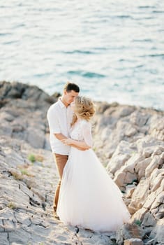 Groom hugs bride in a white lace dress on the rocks by the sea. High quality photo