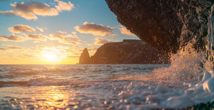 Sea sunset waves with foam. Waves run on pebbles and rock from volcanic basalt. Water breaks into splashes and white foam. Against the background of the cape and the sunset sky