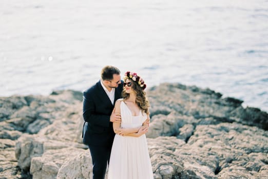 Groom in sunglasses hugs bride in a wreath on the rocks by the sea. High quality photo