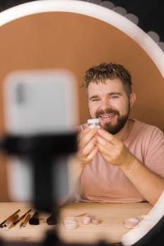 Portrait of happy man beauty blogger sitting in room at table and speaking recommending foundation or decorative cosmetic looking at camera. Blogging and vlog